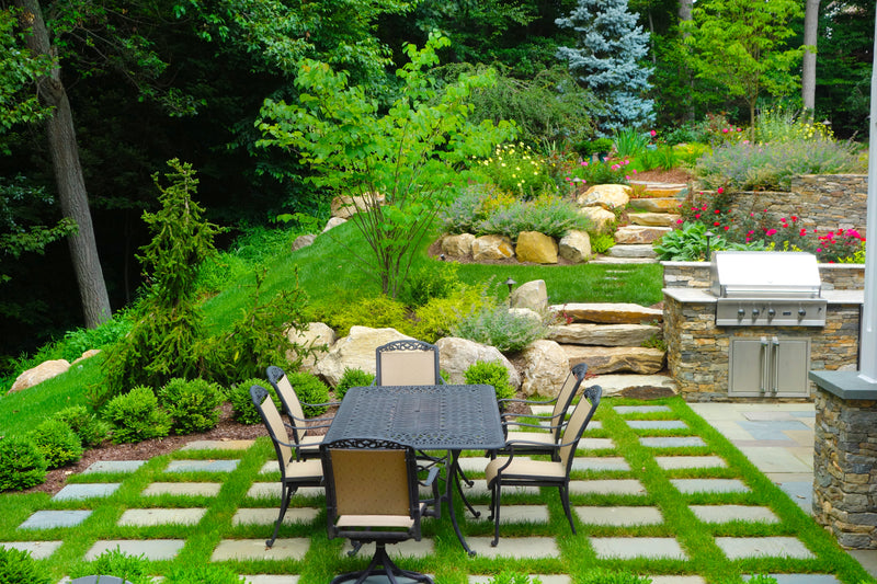 Outdoor patio area with a dining table and chairs, surrounded by greenery and a stone barbecue.